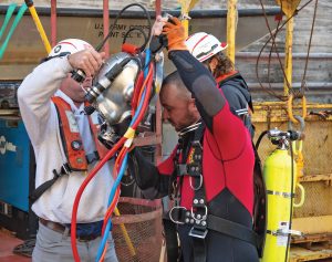 Ty Melton, a lock mechanic with the Nashville Engineer District, dons diving headgear with the help of Matt Williamson, a fellow mechanic at Wilson Lock in Florence, Ala., on October 9. Dive-certified mechanics allow a firsthand look at any underwater maintenance concerns, such as the cracks in the land and river sides of the gates at Wilson that have caused the closure of the main chamber. 